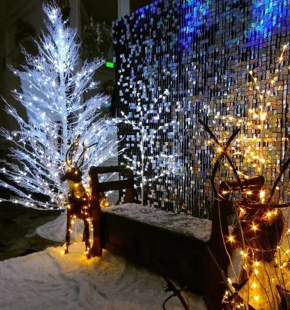 Montaje nocturno con árbol blanco iluminado, renos de luz y muro de lentejuelas